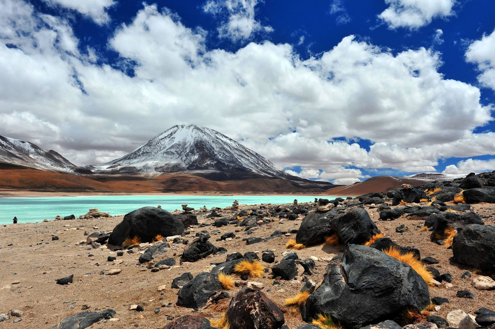 Laguna turquesa con un volcán nevado al fondo y rocas oscuras en primer plano
