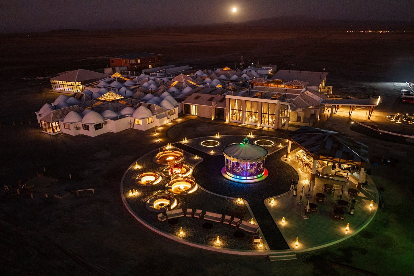 Vue aérienne nocturne d’un hôtel de sel illuminé au bord du salar, avec une place circulaire et un manège