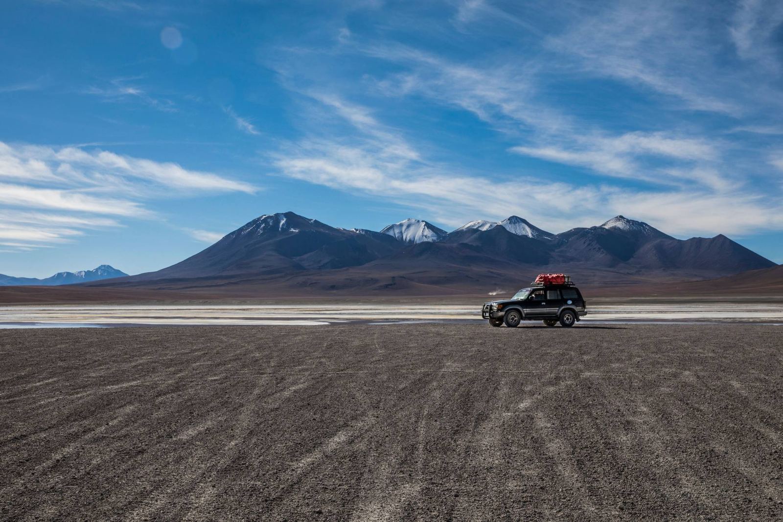 Un 4x4 sur une piste de terre sombre de l’altiplano, avec des volcans et des montagnes au fond