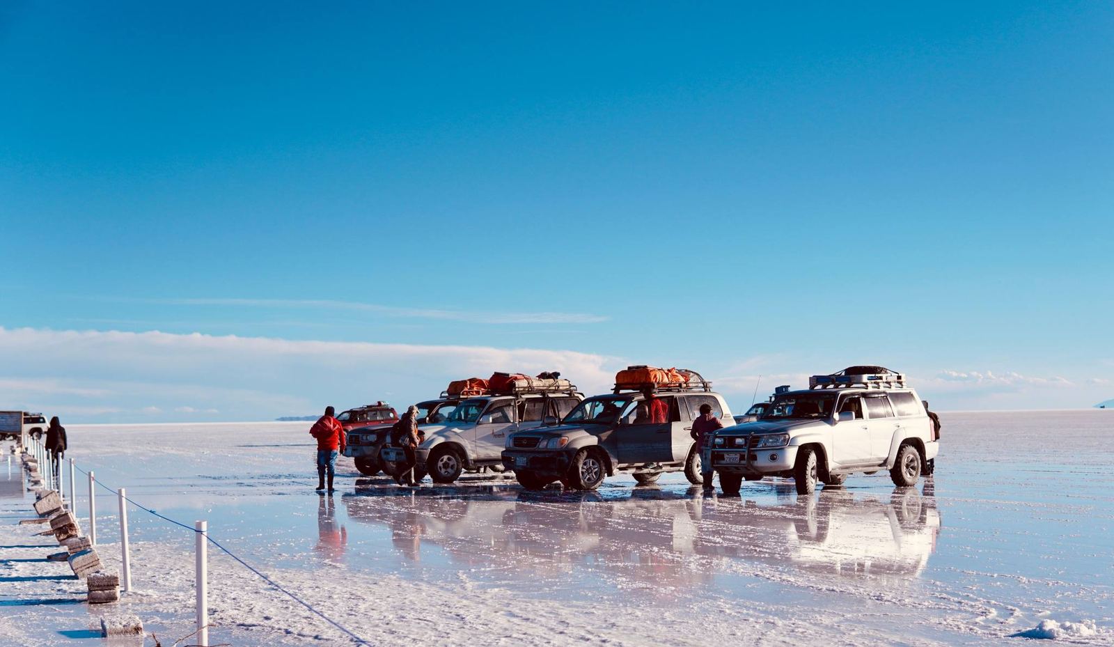 Several 4x4 vehicles with luggage on the roof stopped on the water-covered salt flat, with people next to them