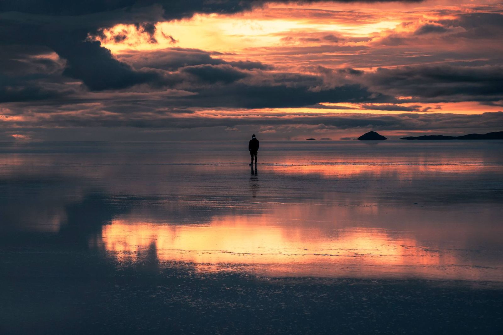 Une personne debout sur le salar recouvert d’eau au coucher du soleil, avec des nuages sombres et un ciel orange reflété