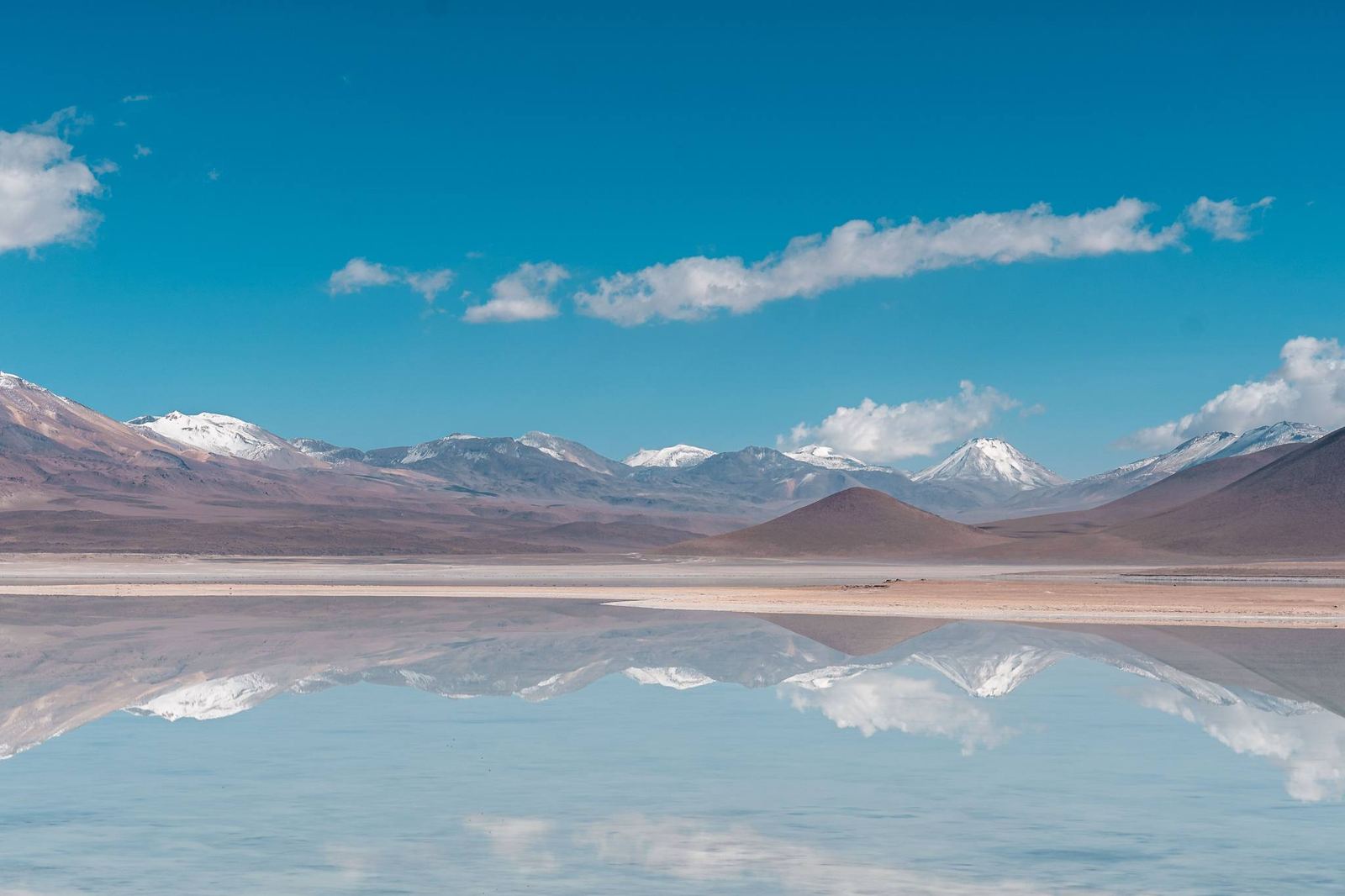 Salar con una fina capa de agua que refleja las montañas con cumbres nevadas y un cielo azul