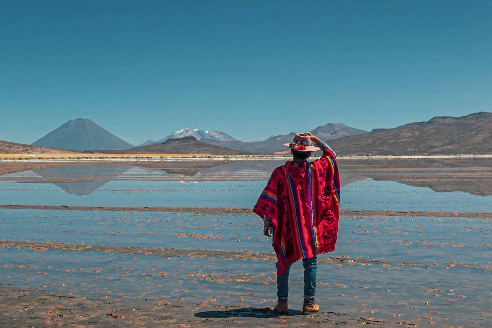 Personne en poncho rouge debout dans l’eau peu profonde du salar, avec un volcan au fond
